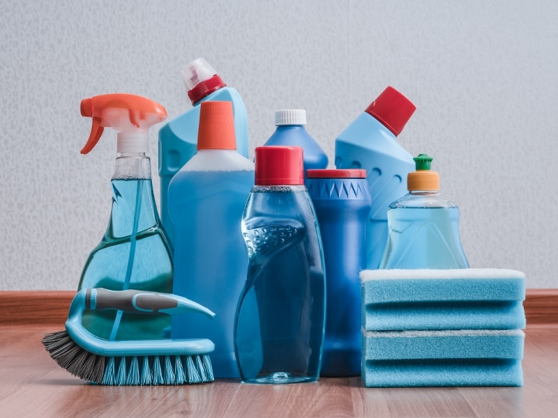 Assorted blue cleaning bottles, brush, spray bottle, and sponges on floor, representing buying cleaning products in bulk